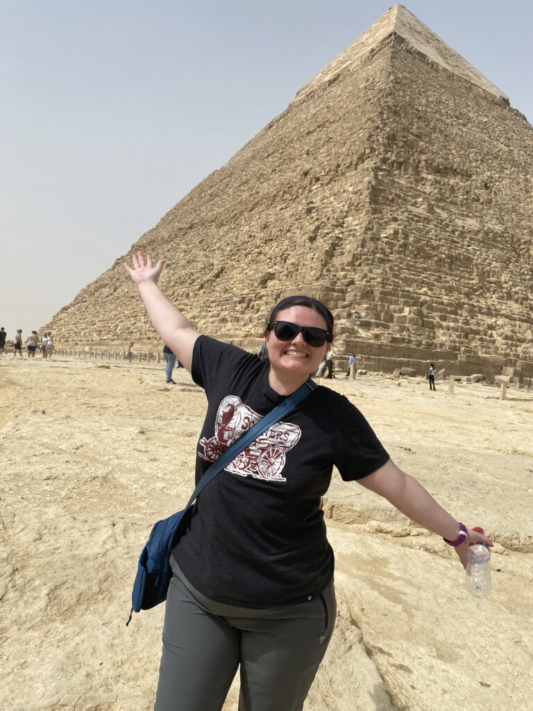 Picture of me (white woman with brown hair in braids in the back wearing black sunglasses). I am standing in front of a pyramid with my arms held out at angles and smiling. 