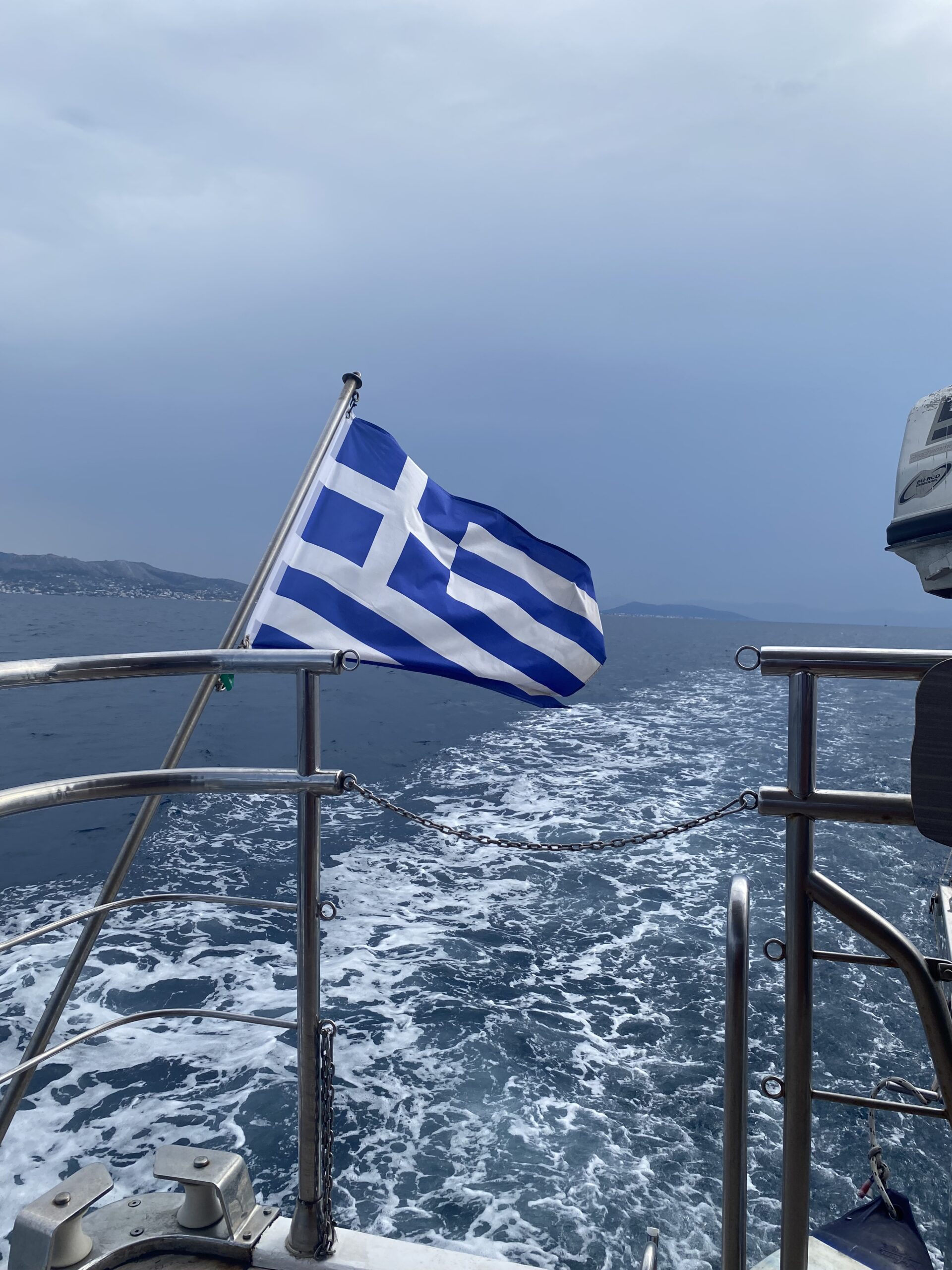 Picture of the back railing of a boat on the ocean with a greek flag blowing in the wind. The Greek flag is Blue and white stripes with a blue square and white plus sign in the top left corner. 