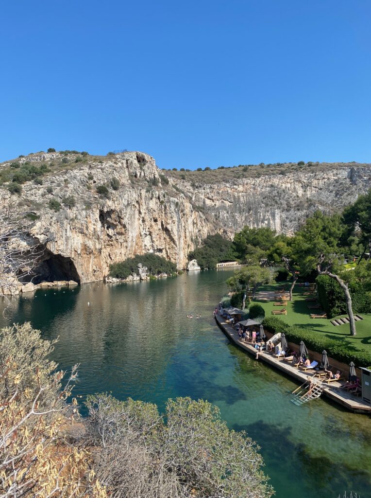 Lake vouliagmeni. the lake is smaller with some rocky hill/cliffs on the left and the lounge area on the right. the lake is a pretty blue green color